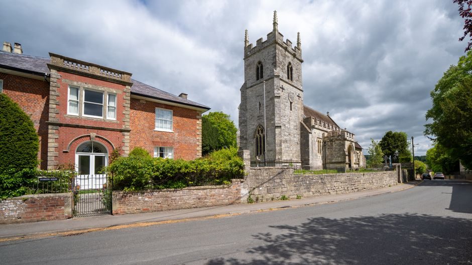 A historic church beside a classic brick house in Codford Saint Peter under a cloudy sky.