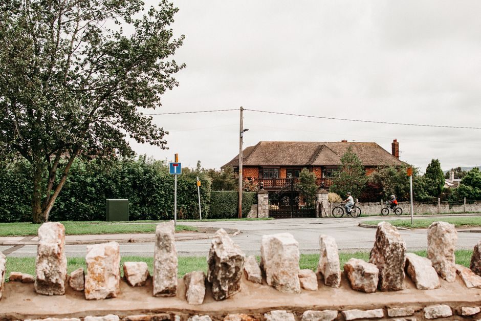 View from behind stone fence on street with bicyclists riding on background of large brick cottage in countryside