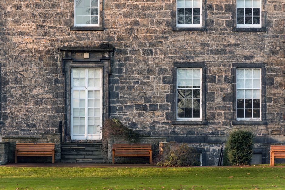 Exterior of old fashioned stone building with windows and door located on grassy lawn with wooden benches in suburb area