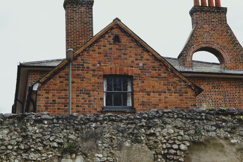 Historic red brick cottage with chimney tops peeking above a weathered stone wall, evoking classic English architecture.